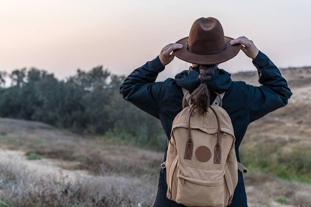 A view of a Caucasian hiker woman wearing a blue jacket, backpack and cowboy hat in the field - Photo, image