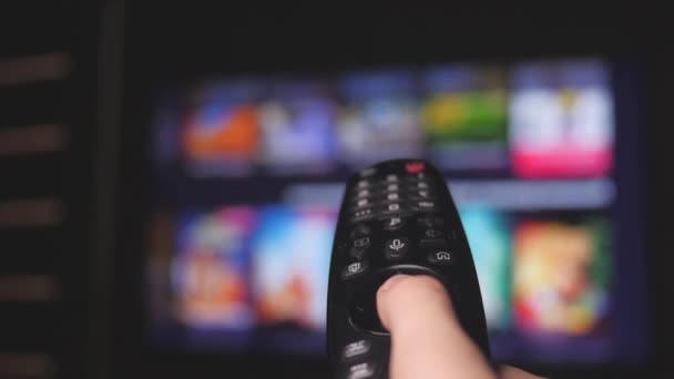 Woman hand selects internet tv channels with remote control, close-up. Person controls TV using a modern remote control. Girl watches smart TV and uses black remote control. Blurry tv scrolls pages - Footage, Video