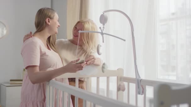 Средний замедленный снимок счастливой молодой беременной женщины с будущей бабушкой, стоящей рядом с детской кроватью, украшенной домашними игрушками-кроватками, улыбающимися и говорящими - Кадры, видео