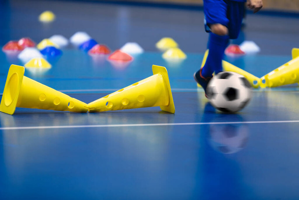 Futsal młody gracz biegnie piłkę na boisku treningowym. Drewniana podłoga do piłki nożnej. Żółte szyszki treningowe na boisku futsalowym. Dzieci szkolące się w futsalu - Zdjęcie, obraz