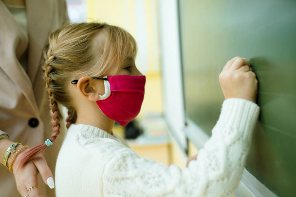A girl with pigtails in a medical mask at the lesson writes in chalk on a blackboard - Photo, Image