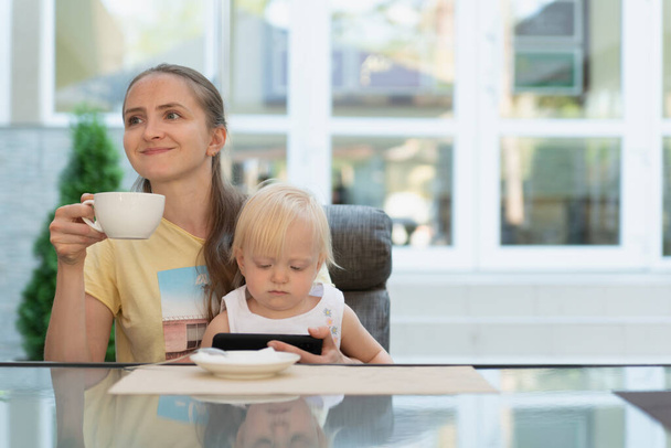 Happy young mom drinks coffee and holds baby in her arms. Kid watching cartoons on phone. - Foto, afbeelding