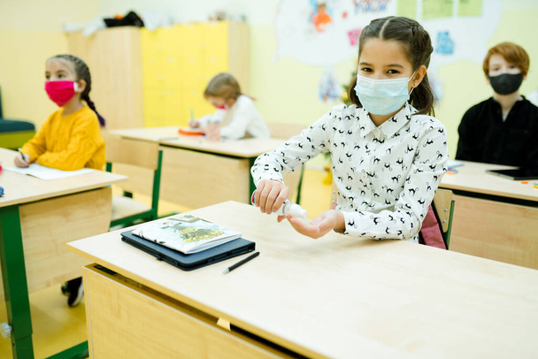 A girl in a white shirt in a medical mask is in the lesson and uses a hand sanitizer - Foto, afbeelding
