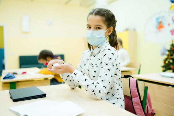 A girl in a white shirt in a medical mask is in the lesson and uses a hand sanitizer - Foto, afbeelding