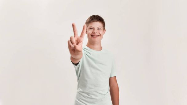 Happy disabled boy with Down syndrome smiling at camera, showing peace sign with one hand while standing isolated over white background - Photo, image