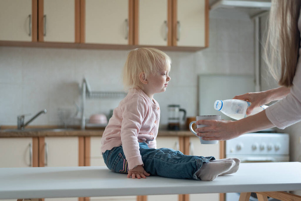 Lapsi istuu keittiössä ja odottaa, että äiti kaataa maitoa mukiin. - Valokuva, kuva