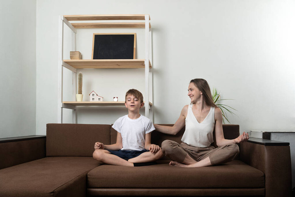 Junge Frau bringt einem Jungen bei, auf Couch zu sitzen und zu meditieren. Yoga zu Hause mit Kindern - Foto, Bild