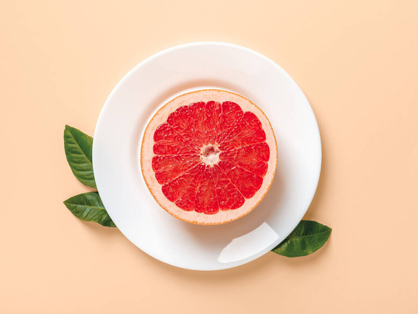 Half of ripe juicy grapefruit on a white plate and few green leaves on a beige background. Slimming diet, body weight control and vitamin healthy eating. Top view. - Zdjęcie, obraz