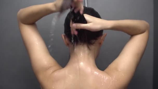 Vue arrière gros plan d'une jeune femme caucasienne aux longs cheveux foncés debout sous la douche dans une salle de bain moderne, se lavant et se rinçant les cheveux - Séquence, vidéo