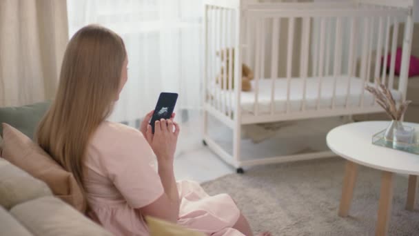Średni widok z tyłu zdjęcia nierozpoznawalnej kobiety siedzącej samotnie w jasnym salonie, patrzącej na zdjęcie USG dziecka z białą kołyską w tle - Materiał filmowy, wideo