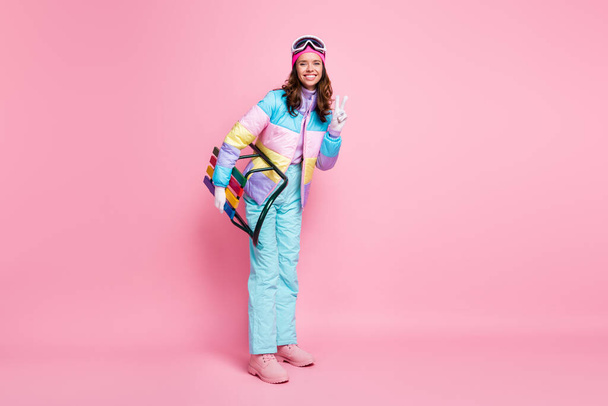 Full length photo of woman skier dressed colorful windbreaker eyeglasses holding sleigh showing v-sign isolated pink color background - Photo, Image
