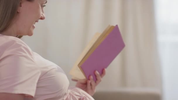 Przechylone zbliżenie młodej kobiety oczekującej rasy kaukaskiej głaskającej brzuch podczas czytania na głos papierowej książki siedzącej na kanapie w jasnym salonie - Materiał filmowy, wideo