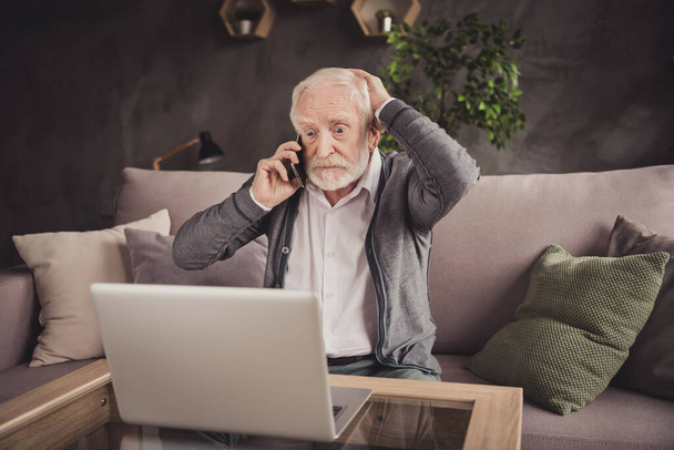 Foto von beeindruckt alten Mann gekleidet weißes Hemd sitzend Sofa suchen modernes Gerät mit Anruf drinnen Wohnung Haus - Foto, Bild