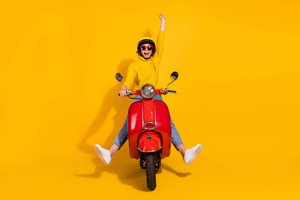 Photo portrait of cheerful girl raising one hand up riding red retro moped isolated on vivid yellow colored background - Фото, изображение