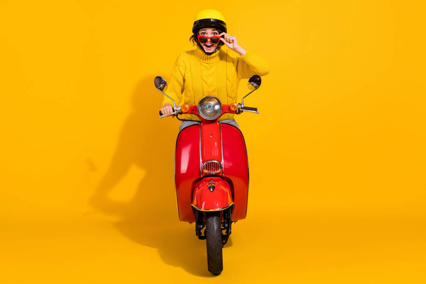 Photo portrait of excited girl pulling down glasses driving red retro scooter isolated on vivid yellow colored background - Valokuva, kuva