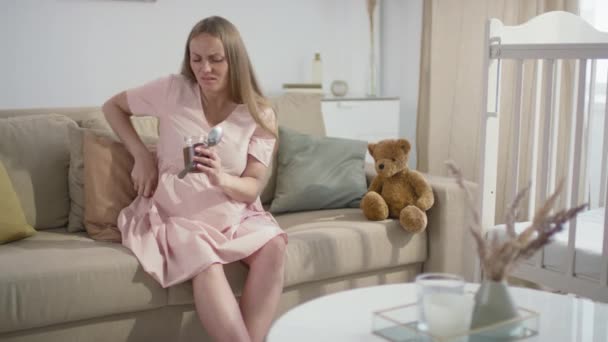 Mittlere Zeitlupenaufnahme einer müden Schwangeren mit einem Glas Schokolade in der Hand, die Rückenschmerzen hat und keine bequeme Pose auf dem Sofa im Wohnzimmer findet - Filmmaterial, Video