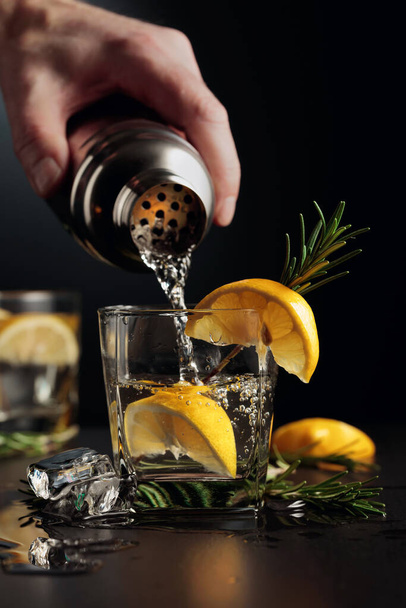 Cocktail Gin ja Tonic sitruunalla ja rosmariinilla. Baarimikko kaataa cocktailin ravistimesta lasiin.. - Valokuva, kuva