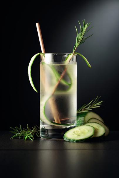 Cold refreshing drink with ice, cucumber, and rosemary. The frozen glass on a black table. - Photo, Image