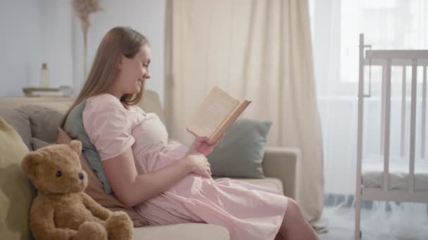 Közepes lassított felvétel egy mosolygó terhes nőről, aki a kanapén ül a nappaliban, Teddy maci mellett, aki hangosan olvas, miközben simogatja a hasát. - Felvétel, videó