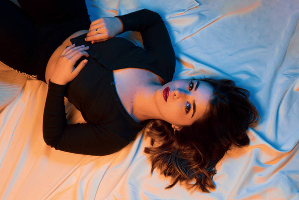 オレンジと青の光でベッドの上に横たわる強烈な視線を持つ美しい若い女性 - 写真・画像