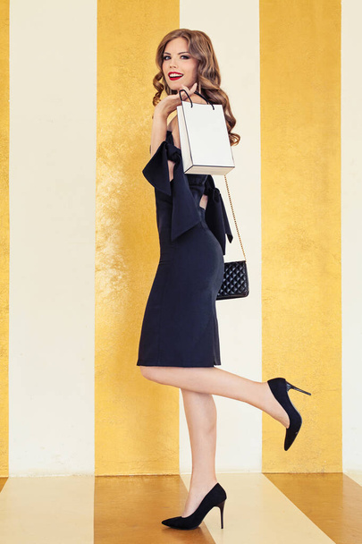Glamoureuze vrouw in zwart cocktail jurk houden shopping tas op gouden achtergrond - Foto, afbeelding