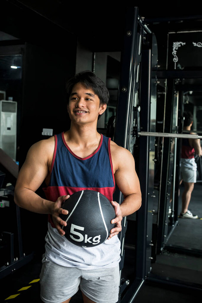 Um jovem asiático confiante e bonito segura uma bola de remédio no ginásio. Um saudável e atlético Gen Z macho. - Foto, Imagem