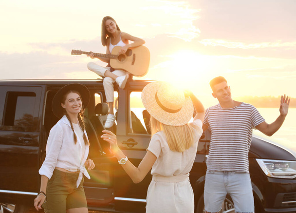 Happy friends having fun near car outdoors at sunset. Summer trip - Zdjęcie, obraz
