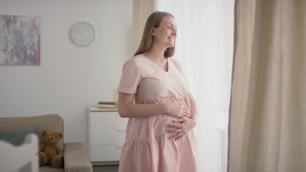 Mediana toma en cámara lenta de hermosa joven embarazada acercándose a la ventana acariciando el vientre y sonriendo disfrutando de la vista - Metraje, vídeo