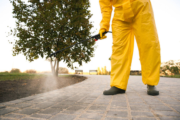 Person in hazmat suit disinfecting pavement in park with sprayer, closeup. Surface treatment during coronavirus pandemic - Fotoğraf, Görsel