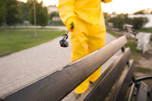 Person in hazmat suit disinfecting bench in park with sprayer, closeup. Surface treatment during coronavirus pandemic - Fotoğraf, Görsel