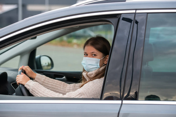 jonge vrouw rijden een auto en het dragen van een beschermend masker tijdens covid 19 pandemie - Foto, afbeelding