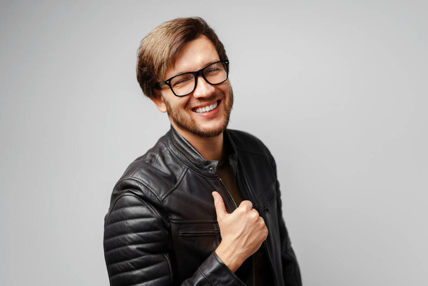 Portrait of a young man in glasses wearing black leather jacket on grey background - Fotó, kép