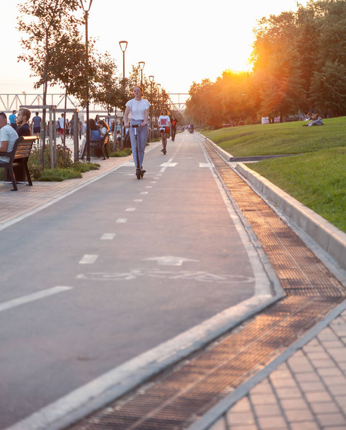 Novoszibirszk, Oroszország - 07.03.2020: Tinédzserek elektromos robogókkal a Mikhailovszkaja töltésen, sokan sétálnak a parkban hétvégenként napnyugtakor egy meleg nyári napon. - Fotó, kép