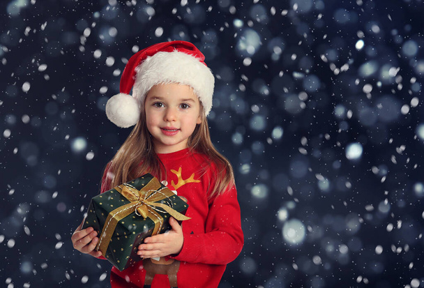 Cute child in Santa hat with Christmas gift under snowfall on dark background - Foto, immagini