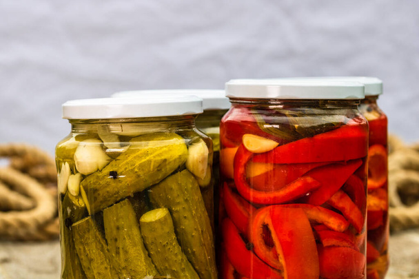Glass jars with pickled red bell peppers and pickled cucumbers (pickles) isolated. Jars with variety of pickled vegetables. Preserved food concept in a rustic composition. - Photo, Image
