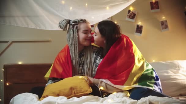 Young lesbian women sitting on the bed covered with LGBT flag - rub their noses and being cute - Footage, Video