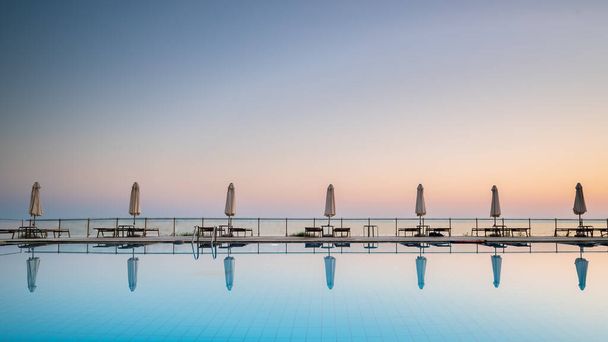 row of sunbeds and sun shades, reflecting in a still swimming pool, as the sun sets, in the Mediterranean - Photo, Image