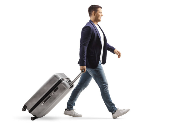 Full length profile shot of a young man smiling and walking with a suitcase isolated on white background - Фото, изображение