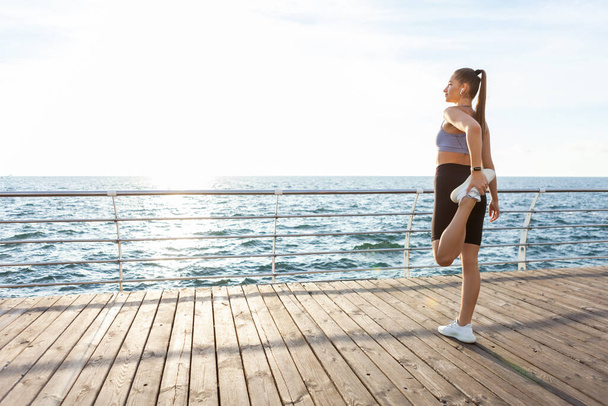 Mujer Slim fit con cuerpo perfecto está practicando estiramiento de piernas antes de entrenar en la playa al amanecer - Foto, Imagen