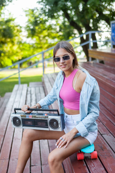 Stylish blonde woman dressed in youth summer clothes listens to music, holding retro boombox player in her hand while sits on a skateboard outdoors - Photo, Image