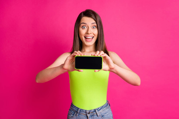 Photo of lovely amazed girl hold telephone presenting touchscreen empty space wear green top isolated vivid pink color background - Фото, зображення
