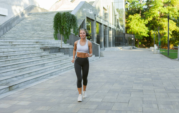 都会の屋外でヘッドフォンで音楽を聴きながら朝のジョギングをするスポーツウェアの若い魅力的な白人ブロンドの女性。朝のワークアウト,健康的なライフスタイル - 写真・画像