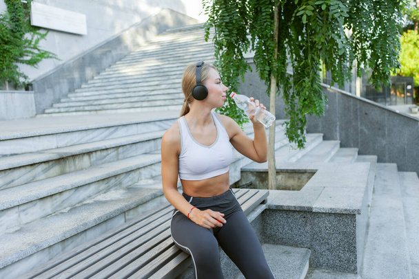 Attractive young fitness blonde woman listens to music on headphones and holds a bottle of water outdoors. Healthy lifestyle concept - Photo, Image