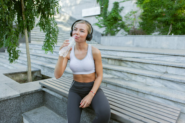Привлекательная молодая блондинка-фитнес слушает музыку в наушниках и пьет воду на открытом воздухе. Понятие здорового образа жизни - Фото, изображение