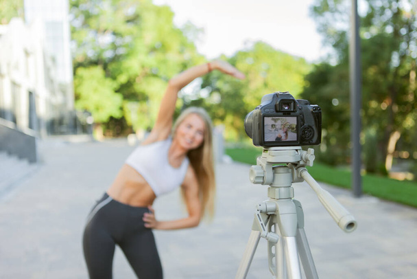 Fitness, sports and video blogging concept. Female sport blogger with camera on tripod recording outdoor fitness training - Photo, Image