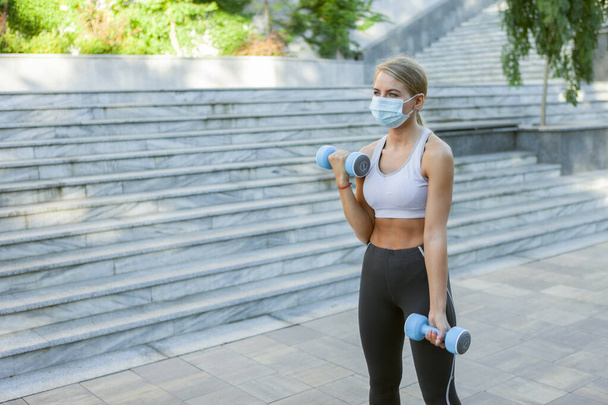 Sportvrouw in sportkleding en medische beschermende gezichtsmasker oefeningen met halters buiten. Fitnesslessen tijdens covid-19 - Foto, afbeelding