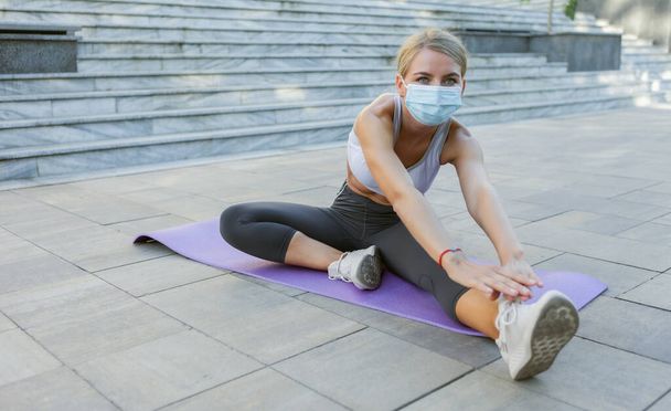 Λεπτή γυναίκα σε φόρμα με ιατρική μάσκα κάνει το πόδι τεντωμένο στο χαλί σε εξωτερικούς χώρους. Γυμναστική άσκηση κατά τη διάρκεια covid-19. - Φωτογραφία, εικόνα