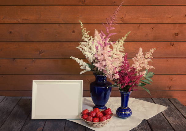 Vermelho, branco e rosa belas flores astilbe em vasos azuis, moldura branca vazia e morango fresco em cesta de vime na mesa de madeira. Vista com espaço de cópia. - Foto, Imagem