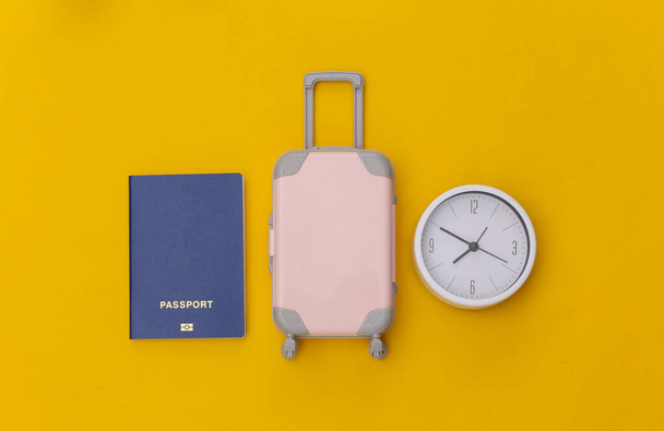 Czas na podróż. Mini plastikowa walizka podróżna i zegar, paszport na żółtym tle. Widok góry. Układy płaskie - Zdjęcie, obraz