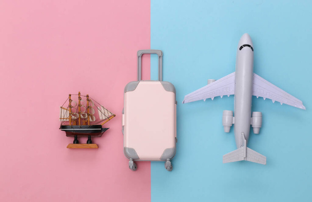 Conceito de viagem. Mini mala de viagem de plástico, navio e avião no fundo pastel azul rosa. Estilo mínimo. Vista superior. Depósito plano - Foto, Imagem